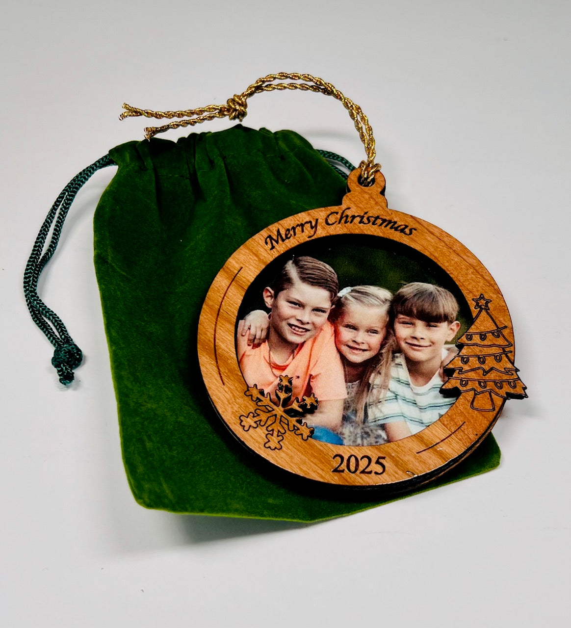Wooden Christmas ornament with photo of three children and 'Merry Christmas 2025' text, on a green velveteen bag.
