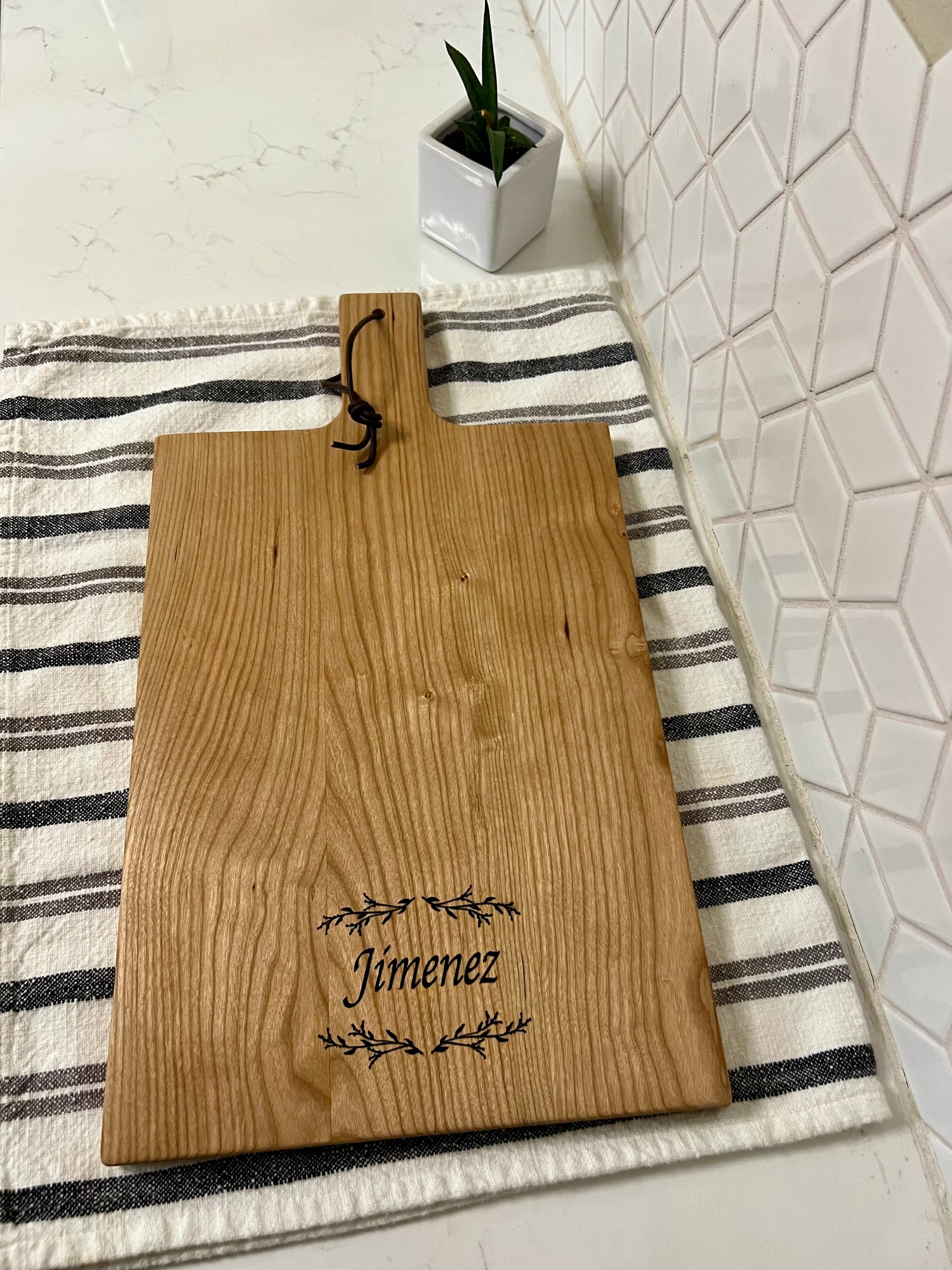 A cherry charcuterie board with a handle and a leather loop, with decorative leaves and Jimenez engraved on it, resting on a floral-patterned cloth next to a potted plant.